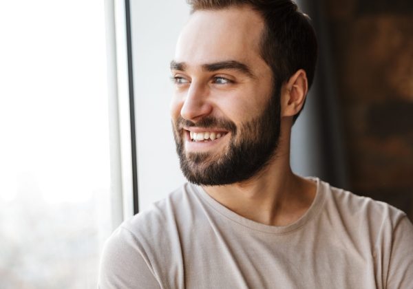Handsome smiling young bearded man standing at the window at home, looking away