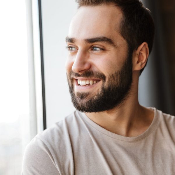 Handsome smiling young bearded man standing at the window at home, looking away
