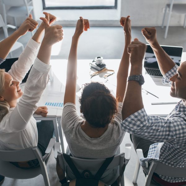 Portrait from back of glad students sitting together at the table and raising hands. Indoor photo of team of freelance specialists having fun after hard work in office.